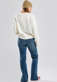 Pull en tricot crème avec des poignets côtelés, associé à un jean bleu évasé. Le mannequin porte un bonnet en tricot marron et des baskets rouges.