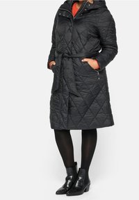 Abrigo largo negro acolchado con cinturón, capucha y bolsillos laterales. Llevado sobre una blusa naranja brillante, combinado con mallas negras y botas de tobillo.