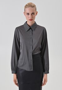Camicia a maniche lunghe bianca e nera con sottili righe diagonali, colletto, chiusura a bottoni e orlo leggermente curvo, abbinata a una gonna nera.