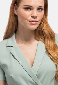 Muntgroene wikkeljurk met korte mouwen, voorzien van een v-hals en een zachte textuur. Gouden ketting met een kleine hanger.