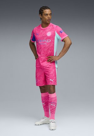 Rosa Fußballtrikot mit strukturiertem Muster, ergänzt durch blaue Akzente und Logos. Umfasst passende Shorts und rosa Socken, getragen mit Fußballschuhen.