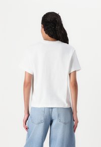 Camiseta blanca de algodón con cuello redondo y mangas cortas, combinada con vaqueros de pierna ancha de mezclilla azul claro, vista desde atrás.