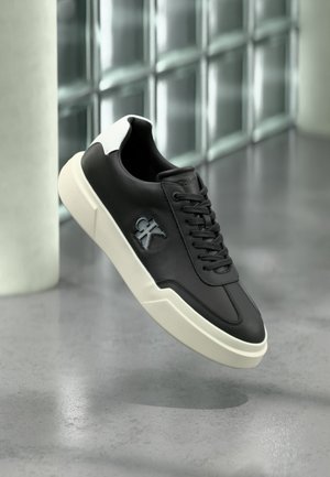 Μαύρο αθλητικό παπούτσι με λευκή σόλα και ταμπελάκι στη φτέρνα, με ανεβασμένο λογότυπο "ck" στην πλευρά, εμφανίζεται σε υπόβαθρο με γκρι γυαλιστερό πάτωμα και γυάλινο τοίχο με κυβάκια.