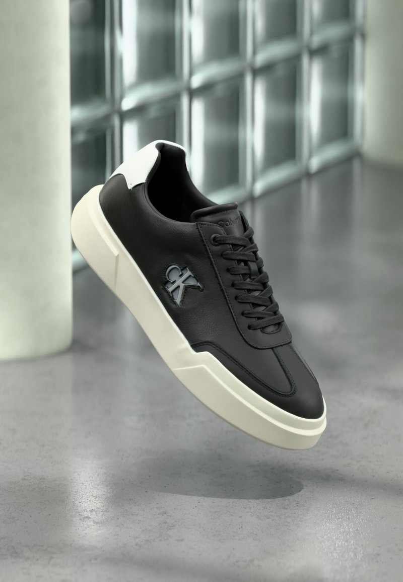 Μαύρο αθλητικό παπούτσι με λευκή σόλα και ταμπελάκι στη φτέρνα, με ανεβασμένο λογότυπο "ck" στην πλευρά, εμφανίζεται σε υπόβαθρο με γκρι γυαλιστερό πάτωμα και γυάλινο τοίχο με κυβάκια.