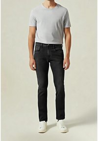 Mężczyzna w jasnoszarej koszuli z krótkim rękawem, czarnych dopasowanych jeansach i białych tenisówkach stoi na tle jednolitego tła.