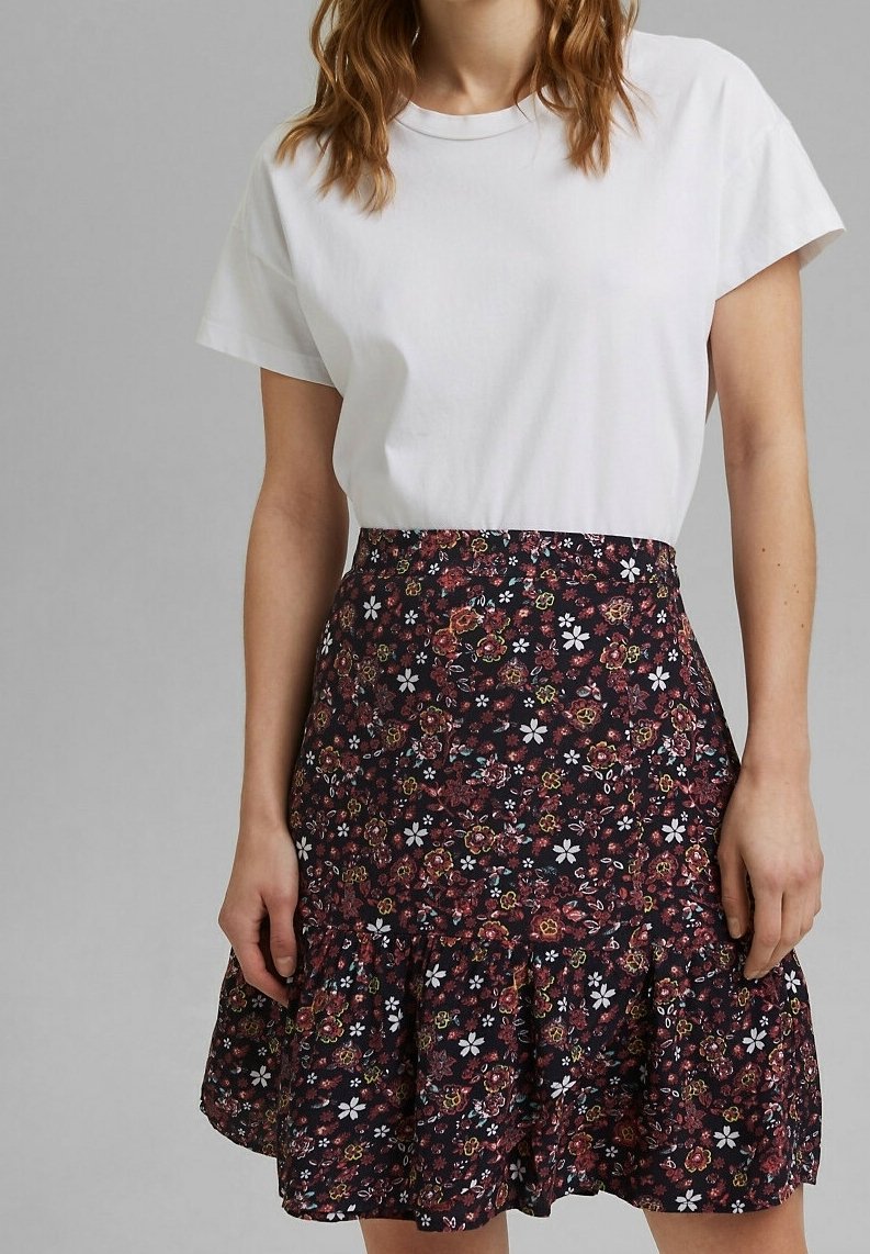 Femme portant un t-shirt blanc à manches courtes rentré dans une jupe foncée avec un petit motif floral coloré et un ourlet volanté.
