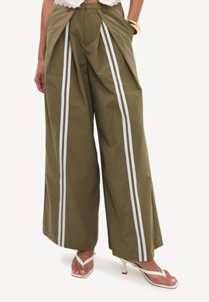 Pantalon large vert olive avec des rayures verticales blanches, porté avec des sandales blanches à talons et un bracelet en or.