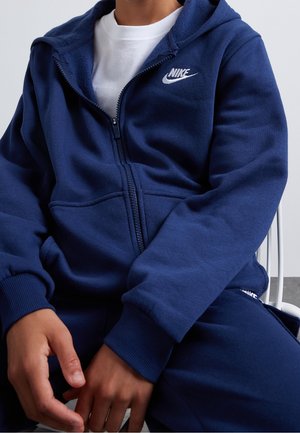 Osoba nosząca granatową bluzę z kapturem i zamkiem marki Nike na białej koszulce, siedząca z rękami spoczywającymi na kolanach.