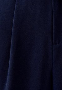 Námorníckom modrom látke s hladkou textúrou, s jemným leskom a subtílnou mäkkosťou. Dizajn zahŕňa drapériu a bočnú kapsu.