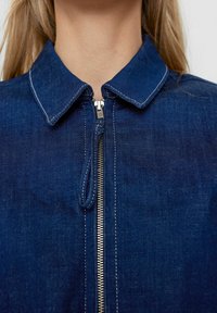 Mörkblå denimjacka med skjortkrage, framzip och vita sömnadsdetaljer. Slät textur med en klassisk design.