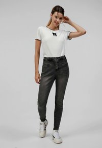 T-shirt blanc à manches courtes avec des finitions noires et un détail en nœud. Jean taille haute gris avec poches avant. Baskets blanches avec des rayures noires.