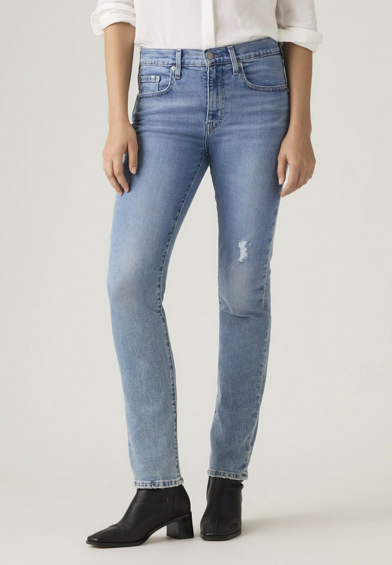 Kvinde iført lyseblå slim-fit jeans med let slid og sorte ankelstøvler med hæl, stående mod en ensfarvet baggrund.