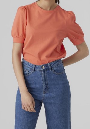 Kvinde iført en koralfarvet t-shirt med pufærmer, stoppet ned i højtaljede blå jeans, med den ene hånd på hoften og den anden afslappet.