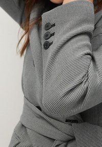 Gros plan sur la manche d'un blazer à motifs pied-de-poule croisé avec trois boutons noirs et une ceinture assortie en tissu nouée à la taille.