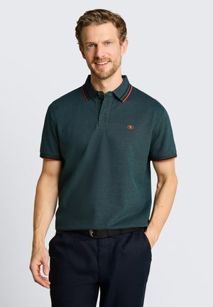 Homme souriant portant un polo vert foncé avec une bordure rouge et un logo, pantalon foncé, debout devant un fond clair uni.
