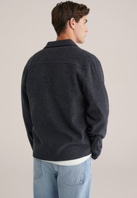 Donkergrijze, gestructureerde, button-up jas met een getailleerde pasvorm, met een kraag en geribde manchetten, gedragen over lichtblauwe jeans.