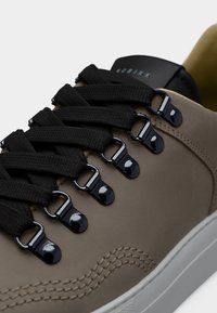 Παντόφλα σε χρώμα ταουπ με μαύρα παχιά κορδόνια περασμένα από γυαλιστερά μεταλλικά άγκιστρα και τριπλή διακοσμητική ραφή κοντά στη λευκή καουτσούκ σόλα.