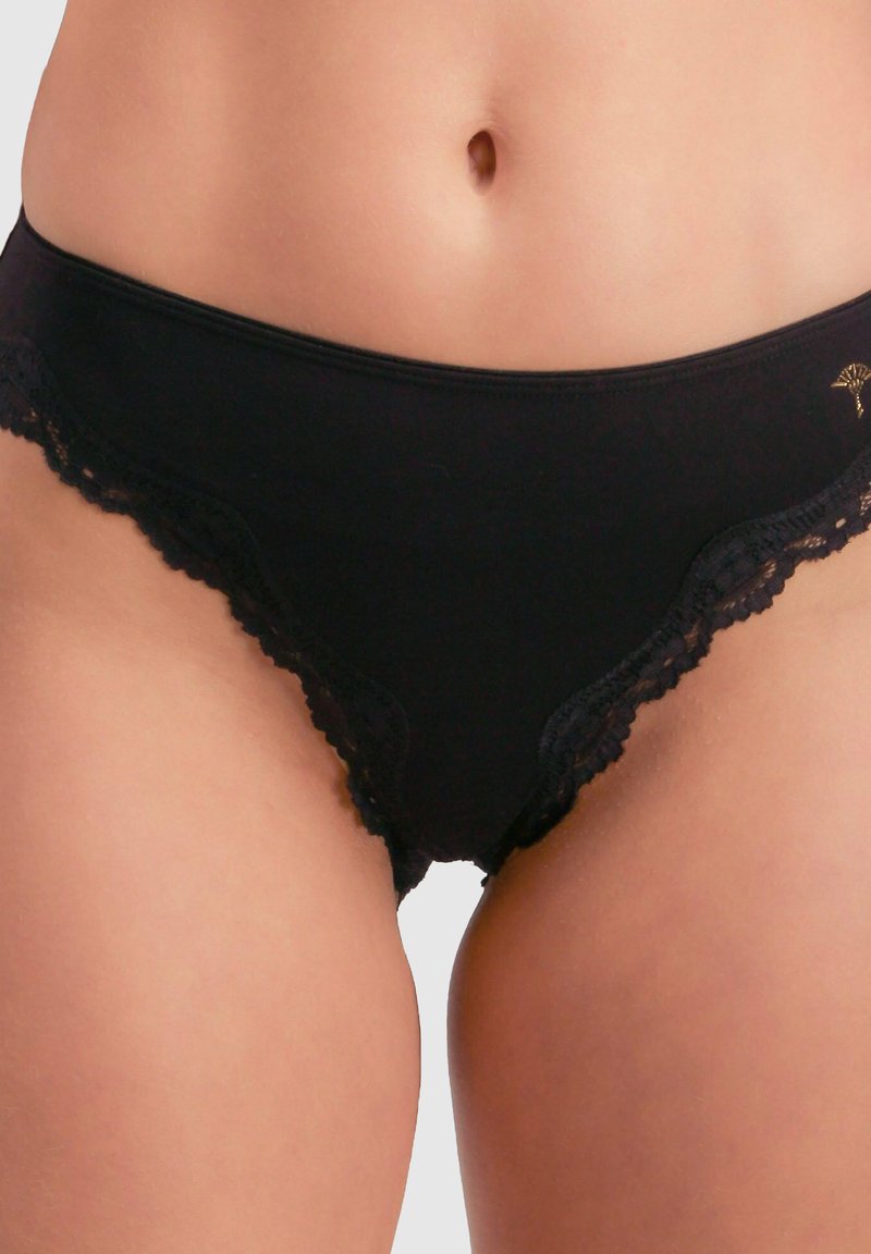 Schwarze Slip mit Spitzenbesatz, die eine glatte Stoffstruktur und ein gewelltes Design an den Rändern aufweisen, mit einem goldenen Logo-Detail auf der Vorderseite.