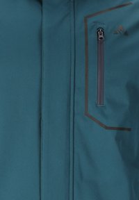 Tealfarbener Jacke von vorne mit vertikaler Reißverschlusstasche, die mit Schwarz umrandet ist, und einem kleinen schwarzen dreieckigen Logo über der Tasche.