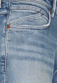 Světle modré džíny z denimu mají klasický pětikapsový design, šité švy a bronzové akcenty. Ošuntělá textura poblíž kapes.