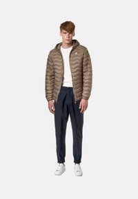 Giubbotto puffer beige con cappuccio, caratterizzato da un motivo a zig zag. Abbinato a pantaloni jogger blu navy e sneakers bianche. Il modello è in piedi con le mani ai lati.