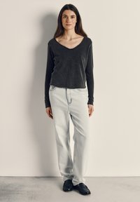 Nainen seisoo yksinkertaista seinää vasten, yllään tumma pitkähihainen paita, vaaleat väljät farkut ja mustat loaferit, katse eteenpäin.