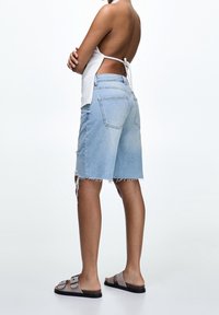 Bermuda shorts in denim blu chiaro con orlo sfrangiato, abbinati a sandali a scivolo marroni. La vista posteriore mostra un design semplice e casual.