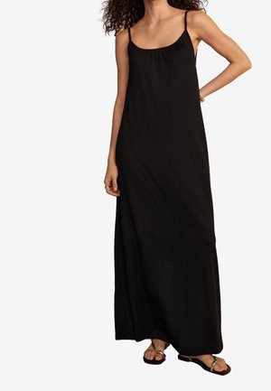 Femme portant une longue robe noire sans manches avec de fines bretelles et des sandales plates marron, debout avec une main sur la hanche.