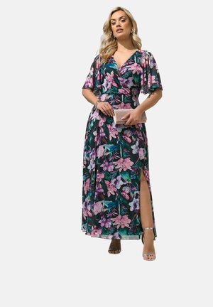 Kvinde iført en lilla og grøn blomstret maxikjole med flagrende ærmer og slids, der holder en beige clutch, stående i højhælede sandaler.