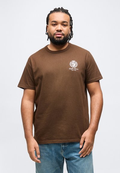 Άνδρας με πλεξούδες στα μαλλιά και γενειάδα, φορώντας καφέ μπλουζάκι Ralph Lauren και μπλε τζιν, στέκεται μπροστά σε απλό ανοιχτό φόντο.