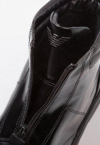 Black ankle boot with a glossy patent finish, featuring a zipper closure and stitched detailing. Logo embossed at the top.