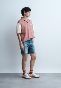 Jeune homme debout, portant des chemises à manches courtes superposées, un short en denim, des chaussettes rayées rouges et des baskets blanches, sur un fond clair uni.