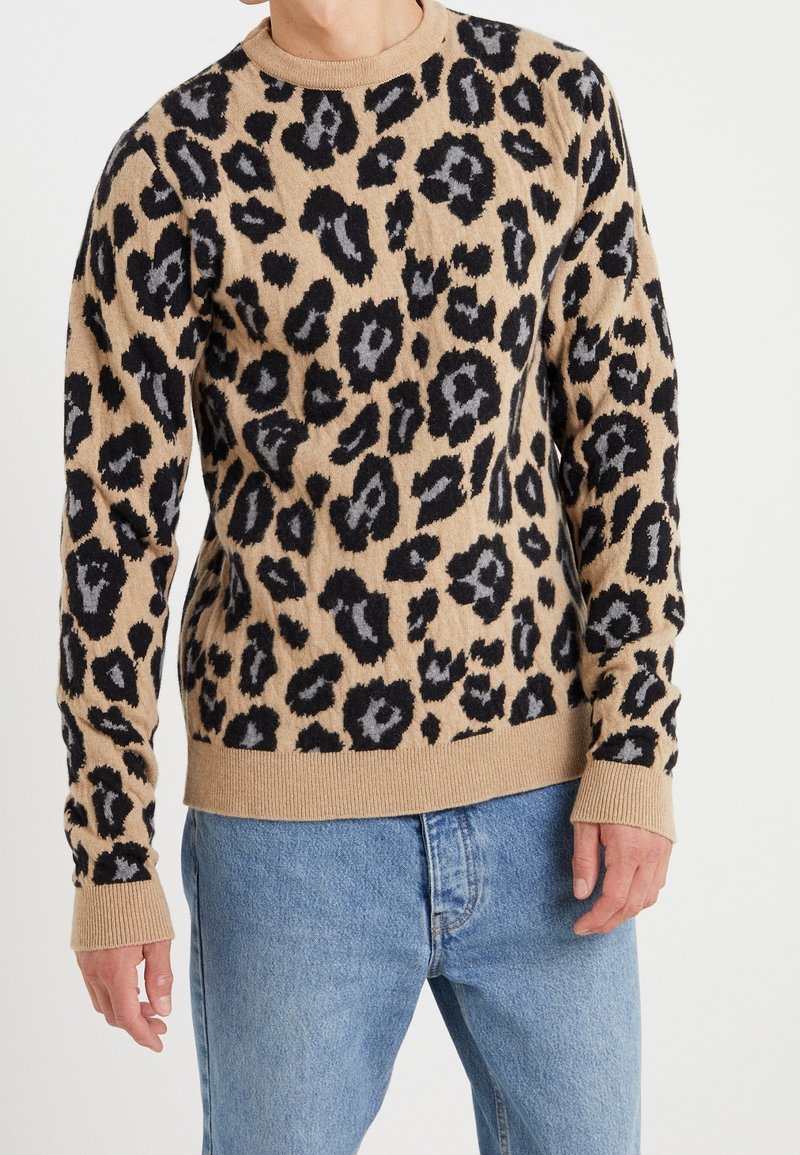 Beige stickad tröja med leopardmönster i svart och grått, med ribbat nacke, ärmar och nederkant. Mjuk textur, figurnära design.