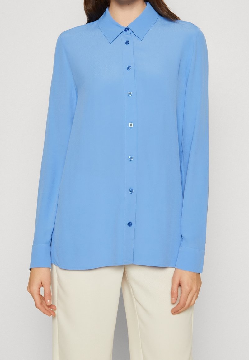 Femme portant une chemise bleu clair à manches longues boutonnée associée à un pantalon écru sur un fond uni.