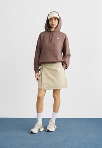 Καφέ φούτερ με κουκούλα, μπεζ φούστα τύπου wrap, λευκές κάλτσες μέχρι τον αστράγαλο, γκρι και λευκά αθλητικά παπούτσια, μπεζ καπέλο με λογότυπο, σε μπλε παρκέ δάπεδο.