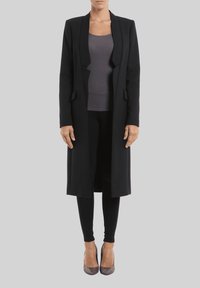 Abrigo largo negro con un diseño a medida, que cuenta con un cuello de chal, dos bolsillos frontales y forrado con un interior suave, llevado sobre una camiseta oscura y leggings.