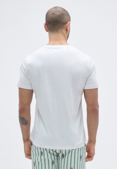 Fehér, rövid ujjú póló kerek nyakkivágással, hátulról látható, zöld-fehér csíkos, mintás nadrággal párosítva.