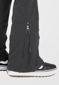 Schwarze, dehnbare Stoffhosen mit einem Reißverschluss am Unterschenkel. Der Stoff ist glatt mit einem leichten Glanz. Kombiniert mit schwarz-weißen Sneakers.