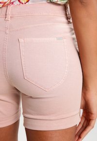 Růžové denimové šortky s ohrnutými manžetami, s jednou zadní kapsou a jemnými švy. Hladký povrch s minimálními hardwarem.
