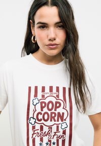 T-shirt blanche avec un graphique de popcorn rayé rouge comportant les inscriptions "POPCORN" et "Frais & Amusant", orné d'accents bleus et de motifs nuage.