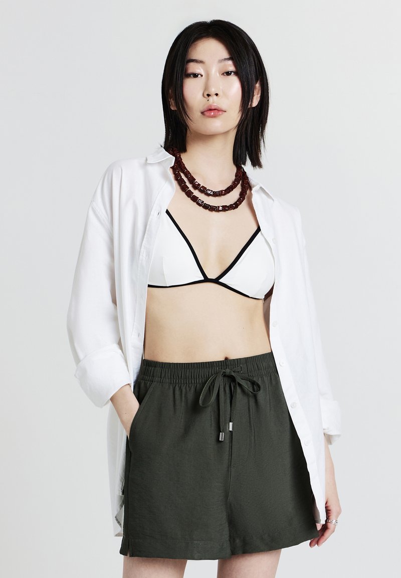 Camicia bianca con bottoni, bralette bianco e nero, pantaloni shorts elastici verde scuro. Collana di perline marrone in polimero, vestibilità casual, stile rilassato.