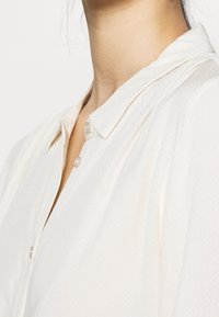 Camicia button-up color crema con superficie texturizzata, tessuto morbido, colletto a punta e bottoni simili a perle, che mette in evidenza un design semplice e pulito.