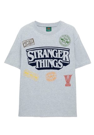 Szürke, rövid ujjú póló, amelyen a "STRANGER THINGS" kék színben látható, különböző színes nyomtatott jelvényekkel és logókkal az elején.