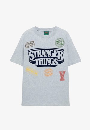 Szürke, rövid ujjú póló, amelyen a "STRANGER THINGS" kék színben látható, különböző színes nyomtatott jelvényekkel és logókkal az elején.