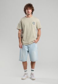 Vaaleanbeige puuvilla t-paita, jossa on pieni graafinen kuva rinnassa, yhdistettynä vaaleansinisiin farkkushortseihin, valkoisiin lenkkareihin ja vihreäraidallisiin sukkiin.