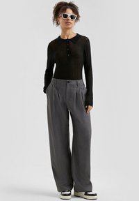 Zwarte lange mouwen blouse met kraag en knopen, gecombineerd met grijze plissé wijde broek. Gedragen met zwart-witte schoenen en witte zonnebril.