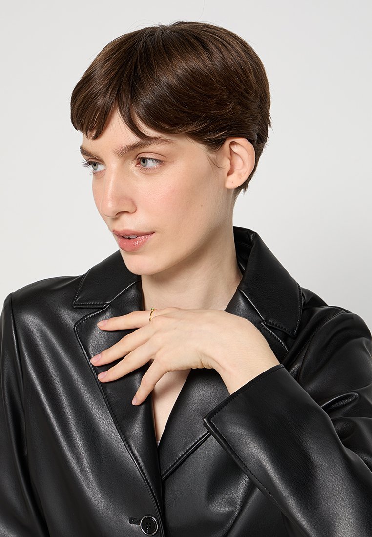 Schwarze Kunstleder-Blazer mit großem Kragen und Knopfverschluss. Das Model posiert mit einer Hand am Kinn und zeigt die glatte Textur sowie das elegante Design.