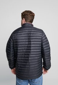 Svart vadderad jacka med horisontell quiltad design, hög krage och kontrasterande röd kant vid halslinningen. Har elastiska muddar och en avslappnad passform.
