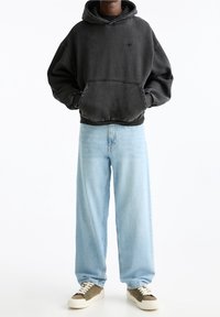 Sudadera con capucha en gris oscuro con un bolsillo frontal, combinada con jeans holgados de color azul claro y zapatillas de color olivo. Ajuste casual y cómodo.