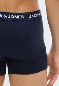 Ανδρικός κορμός που φοράει σκούρο μπλε μποξεράκια με μπλε ζώνη μέσης που φέρει λευκή επιγραφή "JACK & JONES".